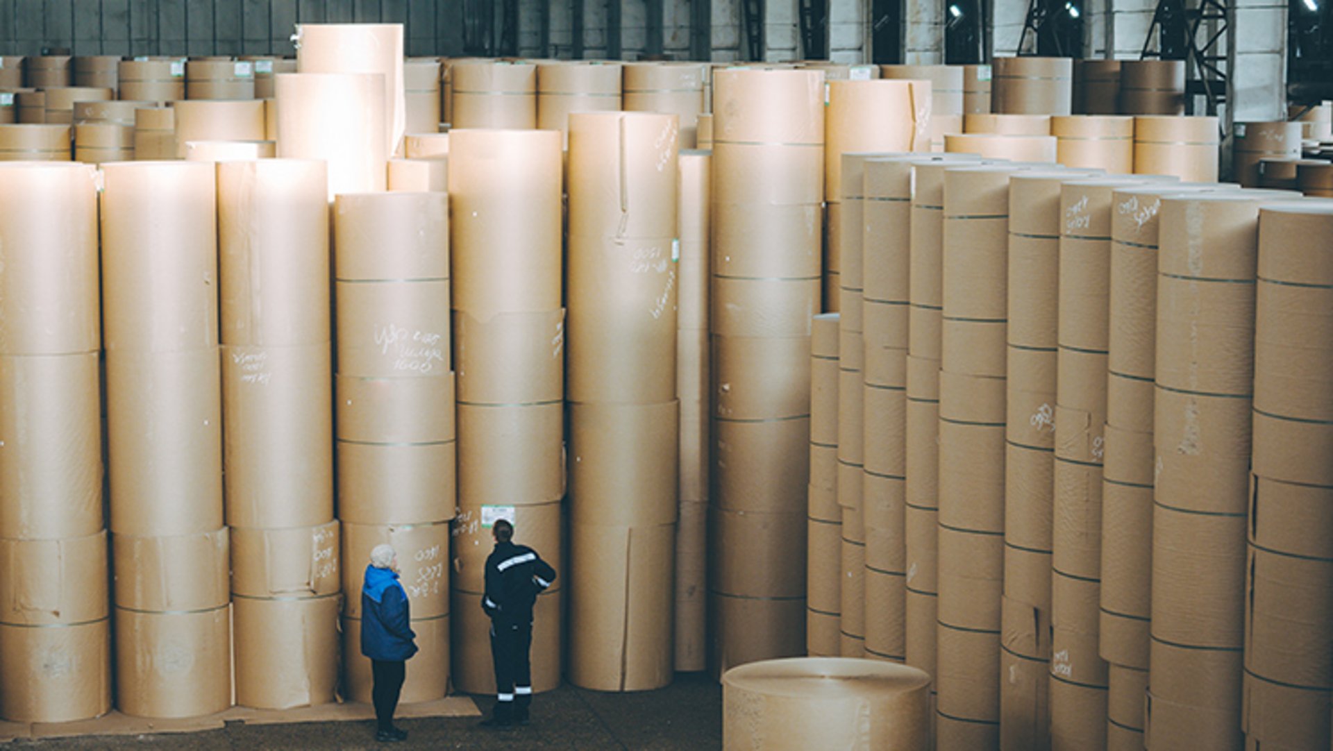 In der Lagerhalle einer Papierfabrik befinden sich zahlreiche Stapel von bis zu sieben Rollen Wellpappenrohpapier, die von zwei Entscheidern begutachtet werden.