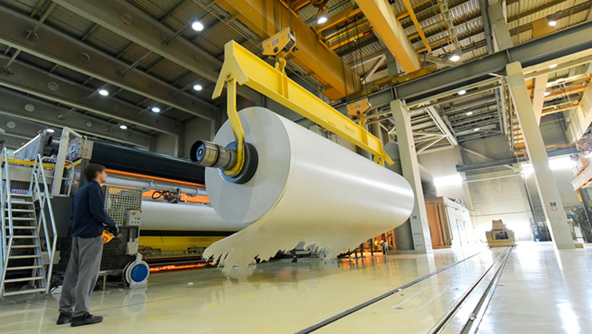 Ein Mitarbeiter einer Papierfabrik steuert in der Werkshalle einen Kran, an dem eine meterbreite und dicke Papierrolle hängt.